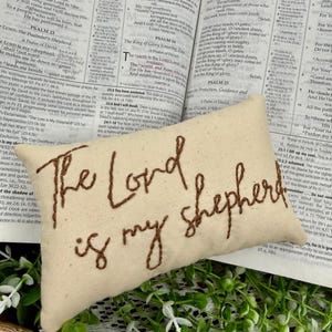 Op de afbeelding: Een beige kussen met de geborduurde zin "The Lord is my shepherd" in bruin garen. Het kussen ligt op een open boek, waarschijnlijk een bijbel, met tekst zichtbaar.