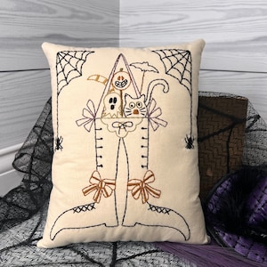May include: Cream-colored Halloween-themed pillow with embroidered black outlines. The design features a witch's boot with a pumpkin, ghost, and cat. Spiderwebs and spiders are embroidered on the sides. Brown and purple accents add to the festive look.