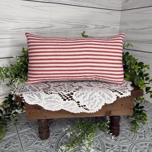 Puede incluir: Un coj&iacute;n rectangular con rayas horizontales rojas y blancas. Se encuentra sobre una peque&ntilde;a mesa de madera cubierta con un mantel de encaje blanco. La vegetaci&oacute;n verde rodea la mesa, creando un ambiente r&uacute;stico.