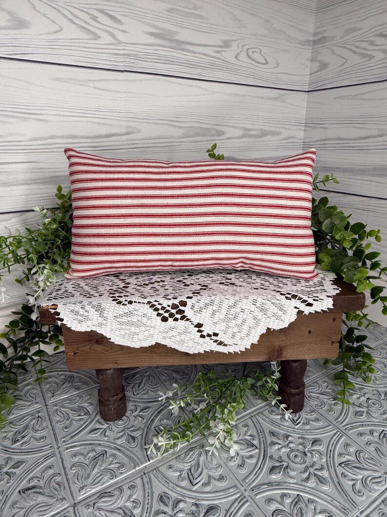 Puede incluir: Un coj&iacute;n rectangular con rayas horizontales rojas y blancas. Se encuentra sobre una peque&ntilde;a mesa de madera cubierta con un mantel de encaje blanco. La vegetaci&oacute;n verde rodea la mesa, creando un ambiente r&uacute;stico.