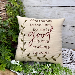 May include: Cream-colored square pillow with embroidered text: "Give thanks to the Lord, for He is good, His love endures forever!" The text is in dark red script. Green embroidered leaves frame the text. Decorative home decor.