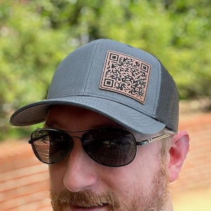 May include: A person wearing a gray and black trucker hat with a brown leather patch featuring a QR code. The person is wearing sunglasses and has a beard.