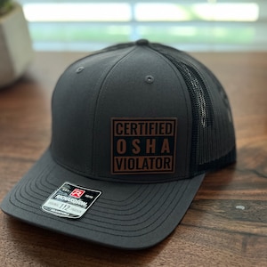 May include: A dark gray trucker hat with a black mesh back. The hat has a brown leather patch with the text "CERTIFIED OSHA VIOLATOR" in white.