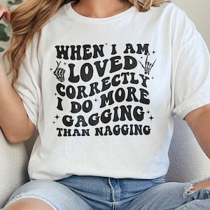 May include: White t-shirt with the text "WHEN I AM LOVED CORRECTLY I DO MORE GAGGING THAN NAGGING" in black. The shirt has a retro font and a skeleton hand graphic. The shirt is worn with blue jeans.