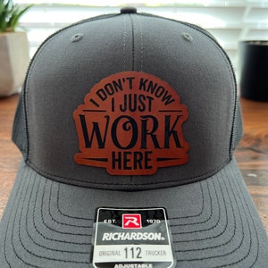 May include: Gray trucker hat with a black mesh back and a brown leather patch that reads "I DON'T KNOW I JUST WORK HERE." The hat has a Richardson 112 Trucker tag attached.