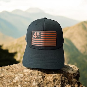 May include: A black trucker hat with a red, white, and blue American flag patch that says "45:7" on it. The hat is sitting on a rock.