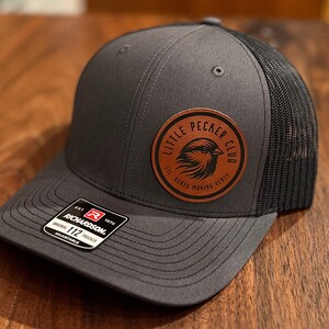 May include: A gray and black trucker hat with a brown leather patch that says "Little Pecker Club" with a bird design. The patch also says "Lil' Birds Making Big Dreams".