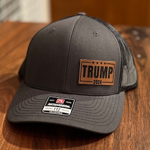 May include: A gray and black trucker hat with a brown leather patch that says "TRUMP 2024". The hat has a black mesh back and a Richardson 112 adjustable strap.
