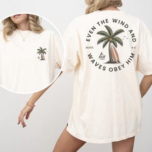 May include: Cream-colored t-shirt featuring a palm tree graphic on the front and back. The back design includes a palm tree, surfboard, and the text "Even the wind and waves obey him" in a circular arrangement. A small palm tree graphic is on the front.