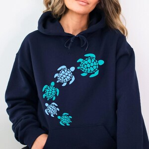 K&ouml;nnte beinhalten: Ein marineblauer Kapuzenpullover mit einem Aufdruck von f&uuml;nf Meeresschildkr&ouml;ten in Blaut&ouml;nen und Gr&uuml;n. Die Schildkr&ouml;ten sind in einer geschwungenen Linie angeordnet, wobei die gr&ouml;&szlig;te Schildkr&ouml;te oben und die kleinste unten ist.