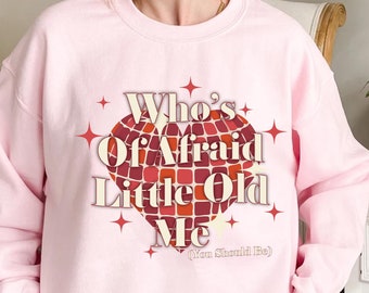 May include: A light pink sweatshirt with the text "Who's Afraid Little Old Me" in a retro font, set within a heart-shaped design of red and orange squares. The sweatshirt has a crew neck.