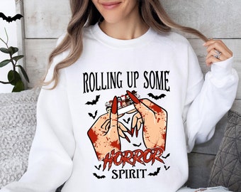 May include: White sweatshirt with a graphic design featuring the text "ROLLING UP SOME HORROR SPIRIT." The design includes bloody hands, bats, and a cigarette with red nails. A Halloween-themed apparel item.