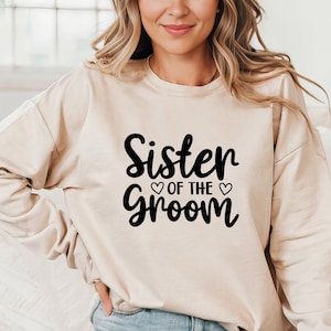 May include: A beige sweatshirt with the text "Sister of the Groom" in black cursive lettering with two small hearts.
