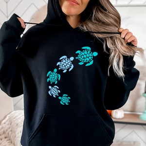 K&ouml;nnte beinhalten: Schwarzer Kapuzenpullover mit einem Aufdruck von f&uuml;nf blauen und gr&uuml;nen Meeresschildkr&ouml;ten, die im Kreis schwimmen.
