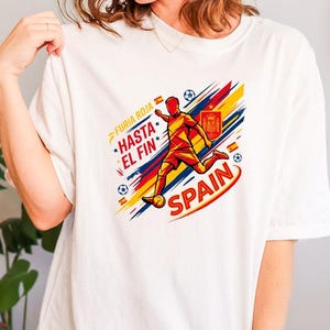 May include: A white t-shirt featuring a graphic of a soccer player in a red and yellow kit kicking a ball. The design includes the text "FURIA ROJA", "HASTA EL FIN", and "SPAIN", along with the Spanish national crest, soccer balls, and red and yellow brush strokes.
