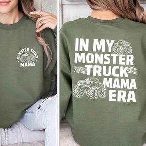 May include: Olive green sweatshirt with white text and graphics. The front features a circular design with a monster truck and the words "Monster Truck Mama." The back displays "In My Monster Truck Mama Era" with monster truck illustrations.