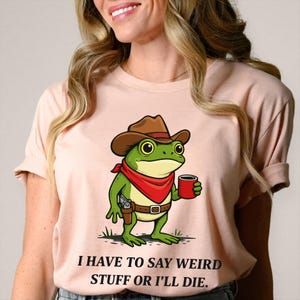 May include: A light pink t-shirt featuring a cartoon frog dressed as a cowboy, wearing a brown hat, red bandana, and holding a red mug. The frog has a gun in a holster. The text on the shirt reads, "I HAVE TO SAY WEIRD STUFF OR I'LL DIE."