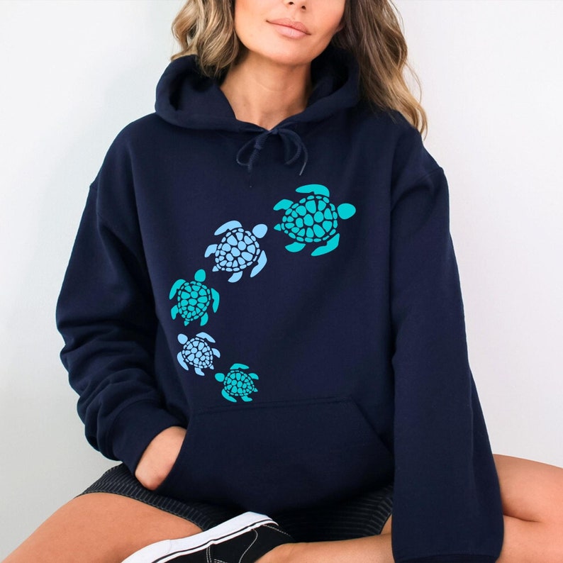 K&ouml;nnte beinhalten: Ein marineblauer Kapuzenpullover mit einem Aufdruck von f&uuml;nf Meeresschildkr&ouml;ten in Blaut&ouml;nen und Gr&uuml;n. Die Schildkr&ouml;ten sind in einer geschwungenen Linie angeordnet, wobei die gr&ouml;&szlig;te Schildkr&ouml;te oben und die kleinste unten ist.