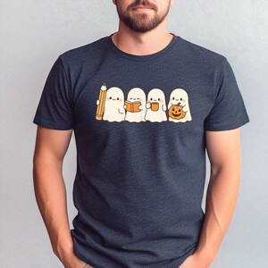 May include: Dark navy blue t-shirt featuring a cute cartoon of four ghosts. One ghost holds a pencil, another a book, the third a mug, and the last a pumpkin. The ghosts are white with black eyes and are smiling.