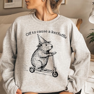 May include: A light gray sweatshirt with the phrase "Off to cause a kerfuffle" printed above a black and white illustration of a beaver wearing a witch hat and riding a scooter. The sweatshirt has a crew neck.