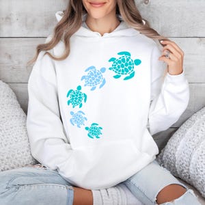 K&ouml;nnte beinhalten: Wei&szlig;er Kapuzenpullover mit einem Aufdruck von f&uuml;nf blauen und t&uuml;rkisfarbenen Meeresschildkr&ouml;ten in einem Kreisdesign.