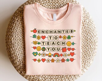 May include: A light pink t-shirt with the words "ENCHANTED TO TEACH YOU" in a playful, colorful design. The design includes hearts, flowers, a rainbow, and a smiling face. The shirt is displayed on a woven, circular mat.