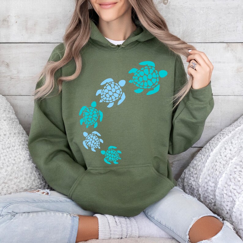 K&ouml;nnte beinhalten: Ein gr&uuml;ner Kapuzenpullover mit einem Design aus f&uuml;nf blauen und t&uuml;rkisfarbenen Meeresschildkr&ouml;ten in einem Kreis auf der Vorderseite.