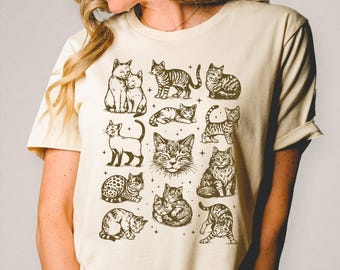 May include: Cream-colored t-shirt featuring a collection of cat illustrations in various poses. The cats are depicted in a vintage style, with detailed line drawings in a dark brown color. The shirt has short sleeves and a relaxed fit.