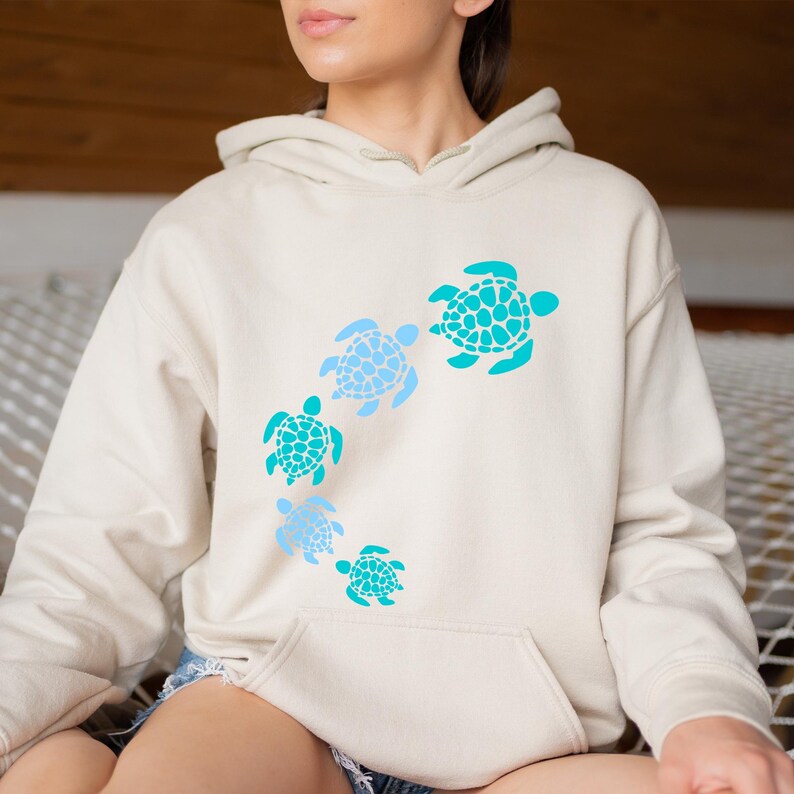 K&ouml;nnte beinhalten: Ein cremefarbener Hoodie mit einem Aufdruck von f&uuml;nf blauen und t&uuml;rkisfarbenen Meeresschildkr&ouml;ten, die im Kreis schwimmen.