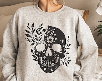 May include: A light gray sweatshirt featuring a black floral skull design. The skull is decorated with leaves and flowers, creating a unique and artistic look. The sweatshirt has a crew neck and long sleeves, perfect for casual wear.