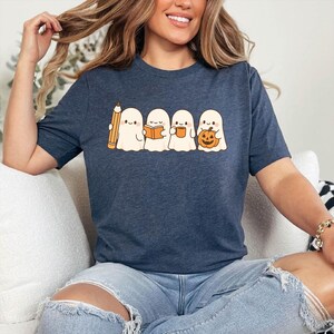 May include: A navy blue t-shirt featuring a graphic of four cartoon ghosts. One ghost holds a pencil, another a book, the third a mug, and the last a jack-o'-lantern. The ghosts are white with black eyes and are on a navy blue background.