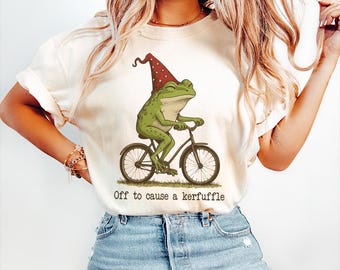 May include: Cream-colored t-shirt featuring a green frog riding a bicycle, wearing a red and white polka-dot hat. The text "Off to cause a kerfuffle" is printed below the frog. The shirt is made of a soft material.
