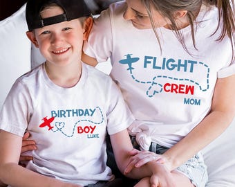 May include: A white t-shirt with a red airplane and the text "Birthday Boy Luke" on it. A white t-shirt with a blue airplane and the text "Flight Crew Mom" on it. A child wearing the "Birthday Boy" shirt and an adult wearing the "Flight Crew" shirt.