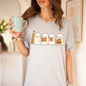 May include: A light gray t-shirt featuring a line of four cartoon ghosts. The ghosts are holding a pencil, a book, a mug, and a jack-o'-lantern. The shirt is a crew neck and has short sleeves.