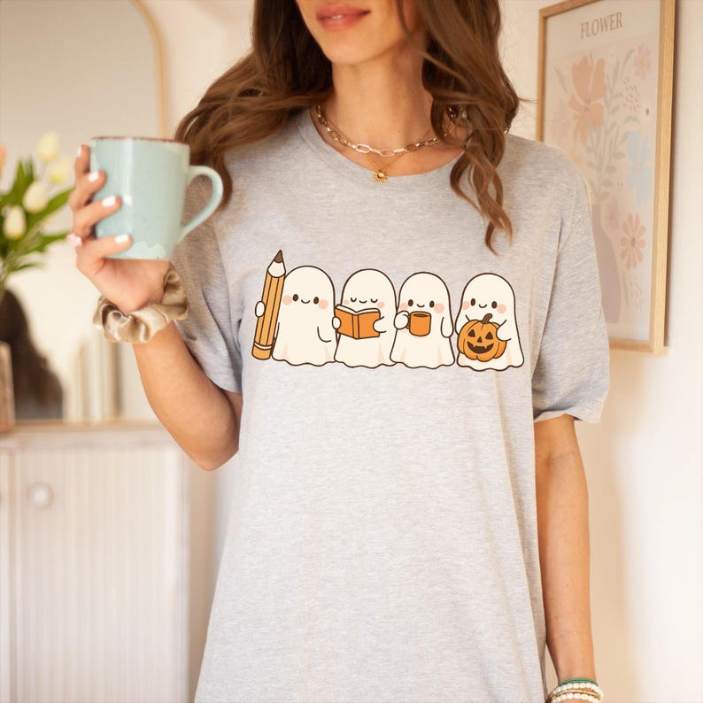 May include: A light gray t-shirt featuring a line of four cartoon ghosts. The ghosts are holding a pencil, a book, a mug, and a jack-o'-lantern. The shirt is a crew neck and has short sleeves.