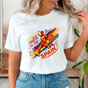 May include: A white short-sleeved t-shirt featuring a graphic of a soccer player in red and yellow. The design includes the text "FURIA ROJA", "HASTA EL FIN", and "SPAIN", along with the Spanish national crest, soccer balls, and diagonal red, yellow, and blue brush strokes.