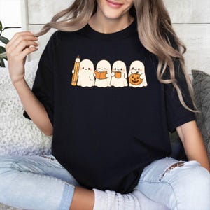 May include: Black t-shirt featuring a line of four cartoon ghosts. The ghosts are holding a pencil, a book, a mug, and a jack-o'-lantern. The ghosts are white with black eyes and are smiling. The pencil is yellow and orange.