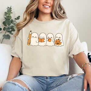 May include: Beige t-shirt featuring a line of cartoon ghosts. One ghost holds a pencil, another a book, a third a mug, and the last a jack-o'-lantern. The design is in a simple, cartoon style, perfect for Halloween.