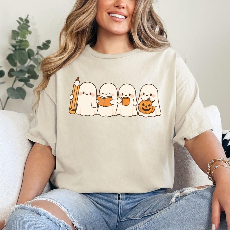 May include: Beige t-shirt featuring a line of cartoon ghosts. One ghost holds a pencil, another a book, a third a mug, and the last a jack-o'-lantern. The design is in a simple, cartoon style, perfect for Halloween.