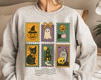May include: A light gray sweatshirt featuring six Halloween-themed postage stamp designs. The stamps depict a jack-o'-lantern, ghost, lollipop, black cat, haunted house, and skull with a tombstone. The designs are in orange, purple, green, and yellow.