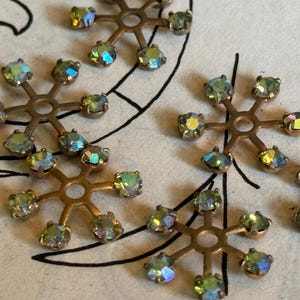 May include: Close-up of several snowflake-shaped embellishments. Each has a bronze-colored metal base with six arms, each holding an iridescent, multi-colored rhinestone. The rhinestones reflect shades of green, blue, and yellow.