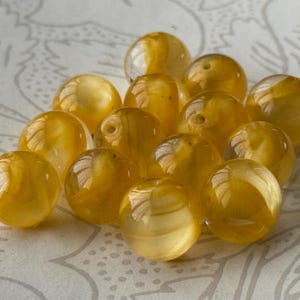 May include: A collection of translucent, golden-yellow beads with a glossy finish. The beads are round and vary slightly in size, with visible internal patterns. The beads are scattered on a patterned, off-white surface.