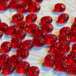 May include: A collection of translucent red beads, each shaped like a strawberry. The beads are scattered across a light-colored surface, with their glossy surfaces reflecting light. The beads are a vibrant red color.