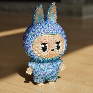 May include: A decorative figurine of a rabbit-like character, adorned with iridescent blue and clear crystals. The figure has large, black eyes and a light beige face. It is standing on a wooden surface.