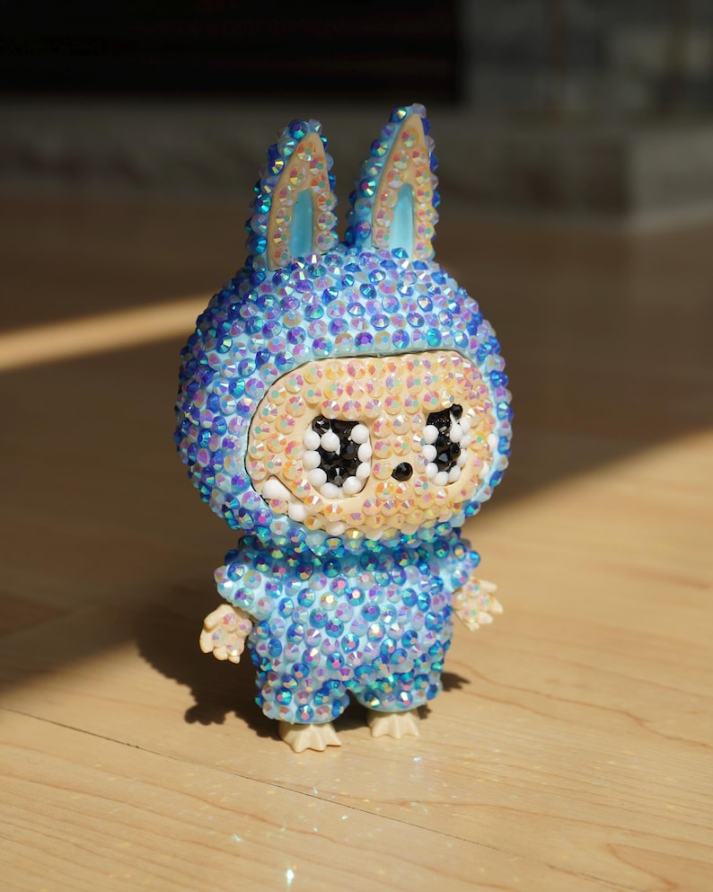 May include: A decorative figurine of a rabbit-like character, adorned with iridescent blue and clear crystals. The figure has large, black eyes and a light beige face. It is standing on a wooden surface.