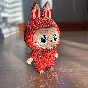 May include: A red and orange figurine of a rabbit-like character, covered in sparkling rhinestones. The figure has large, expressive eyes and small, detailed hands and feet. The background is blurred, highlighting the figurine.