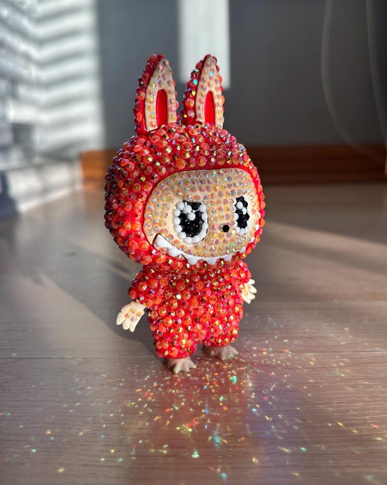 May include: A red and orange figurine of a rabbit-like character, covered in sparkling rhinestones. The figure has large, expressive eyes and small, detailed hands and feet. The background is blurred, highlighting the figurine.