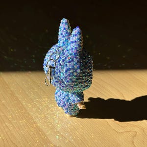 May include: A blue and iridescent bunny-shaped keyring. The keyring is covered in small, sparkling embellishments. The bunny has long ears and a small, rounded body. A silver-coloured keyring and clasp are attached to the top of the bunny.