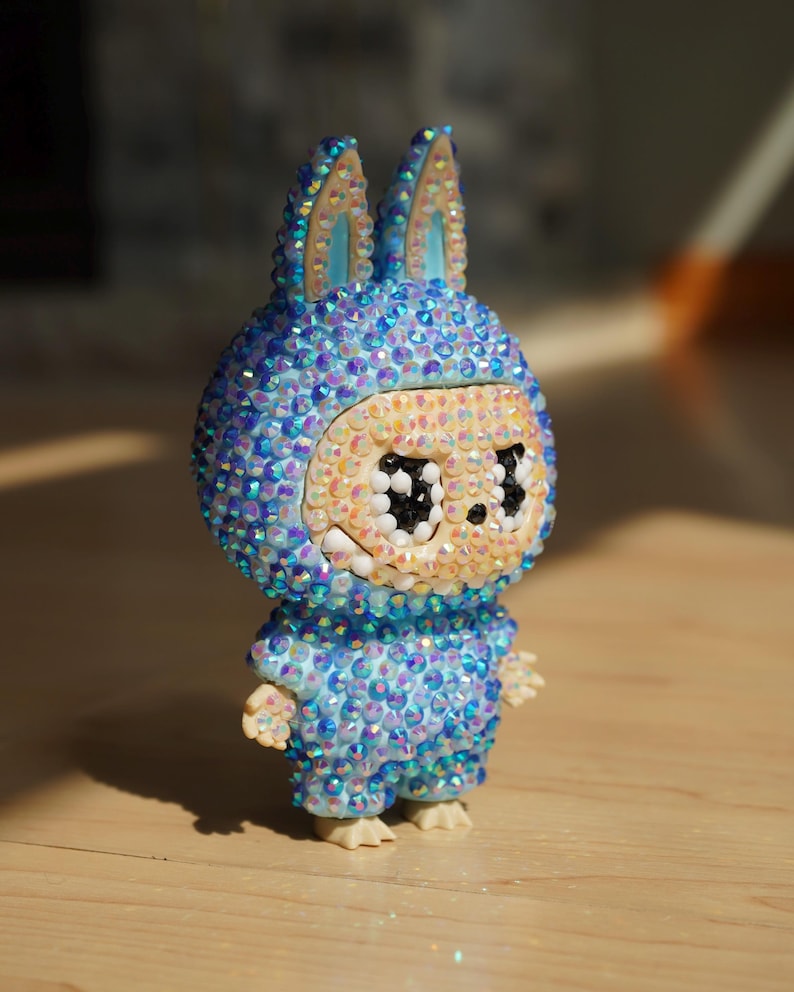 May include: A decorative figurine of a bunny-like character, covered in iridescent blue and clear crystals. The figure has large eyes and wears a matching jumpsuit. The ears are light blue with crystal detailing. The figurine stands on a wooden surface.