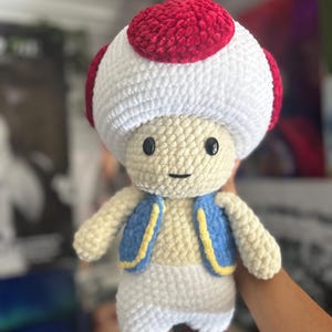 May include: A crochet plush toy shaped like a mushroom character with a large white head topped with a red cap. The figure has black bead eyes, a small stitched mouth, a blue vest with yellow trim, white pants, and brown feet, all made of soft yarn.